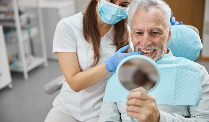 Older man sitting in a dental chair looking in a handheld mirror at his implant supported dentures from Timothy H. Kindt, DDS in Mesa, AZ