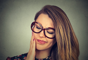  woman with glasses on with sensitive tooth closing her eyes and making a painful expression while touching her face