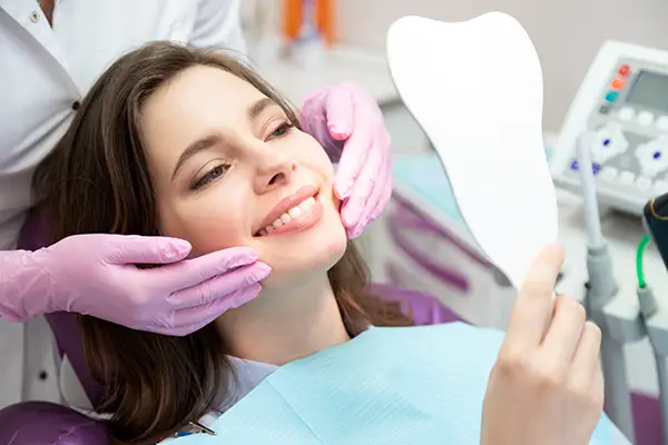 A smiling woman holding a tooth-shaped mirror while a dentist adjusts her face, showcasing her smile makeover results in a clinic.